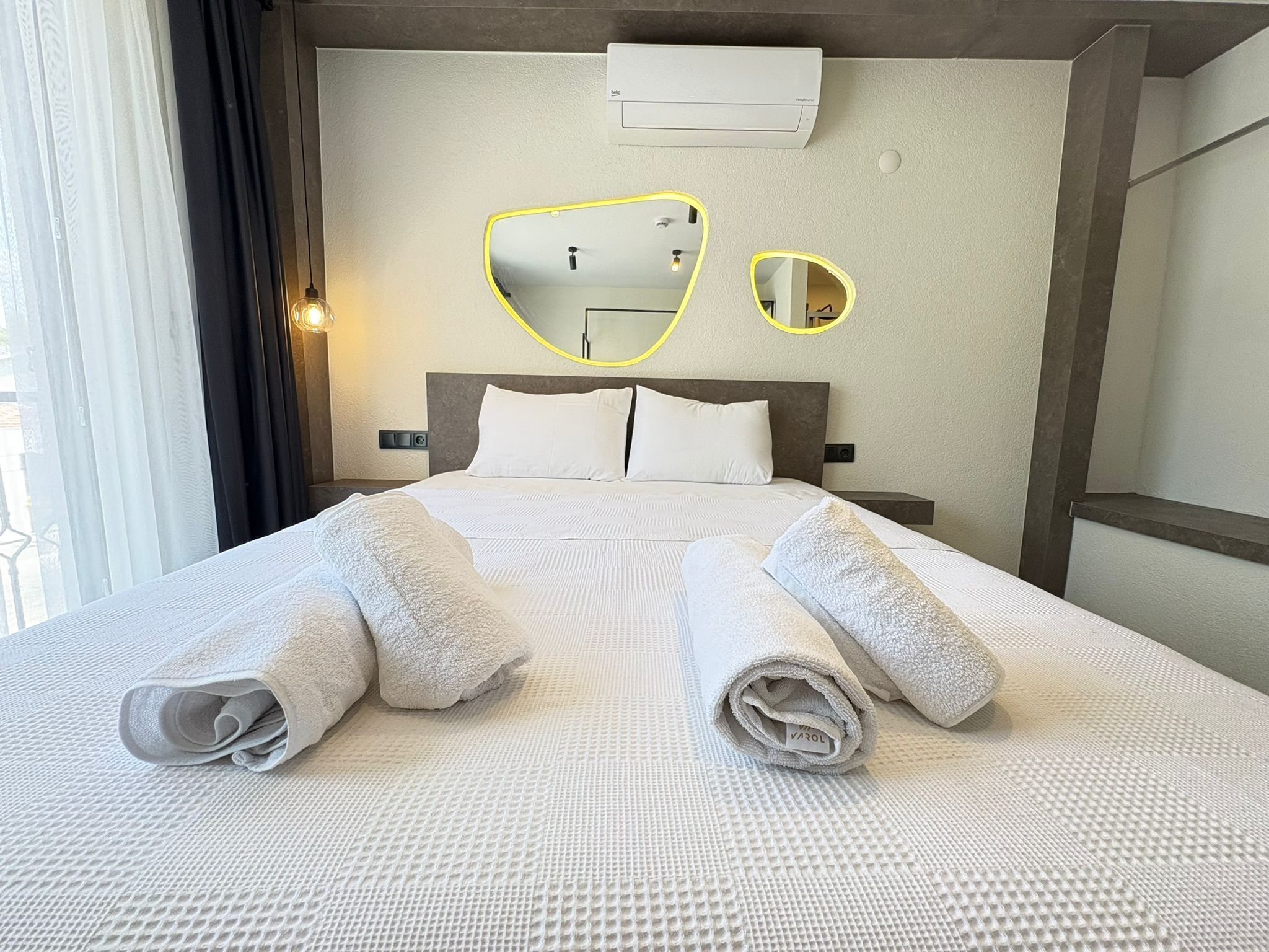 A neatly made bed with two rolled towels, a wall-mounted air conditioner, and a mirror with a yellow frame.
