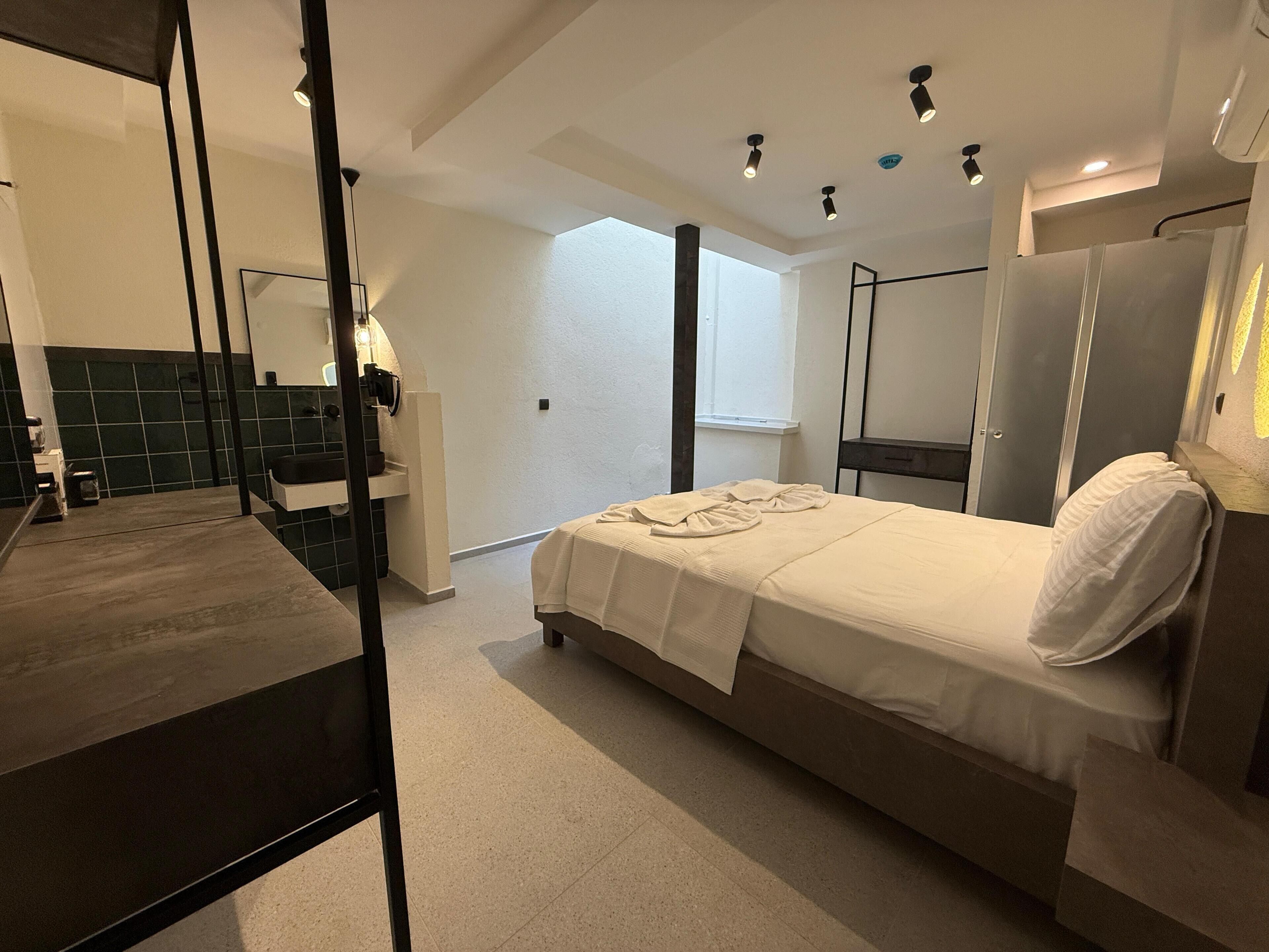 A modern hotel room with a bed, a large mirror, a vanity with a sink, and a window providing natural light.