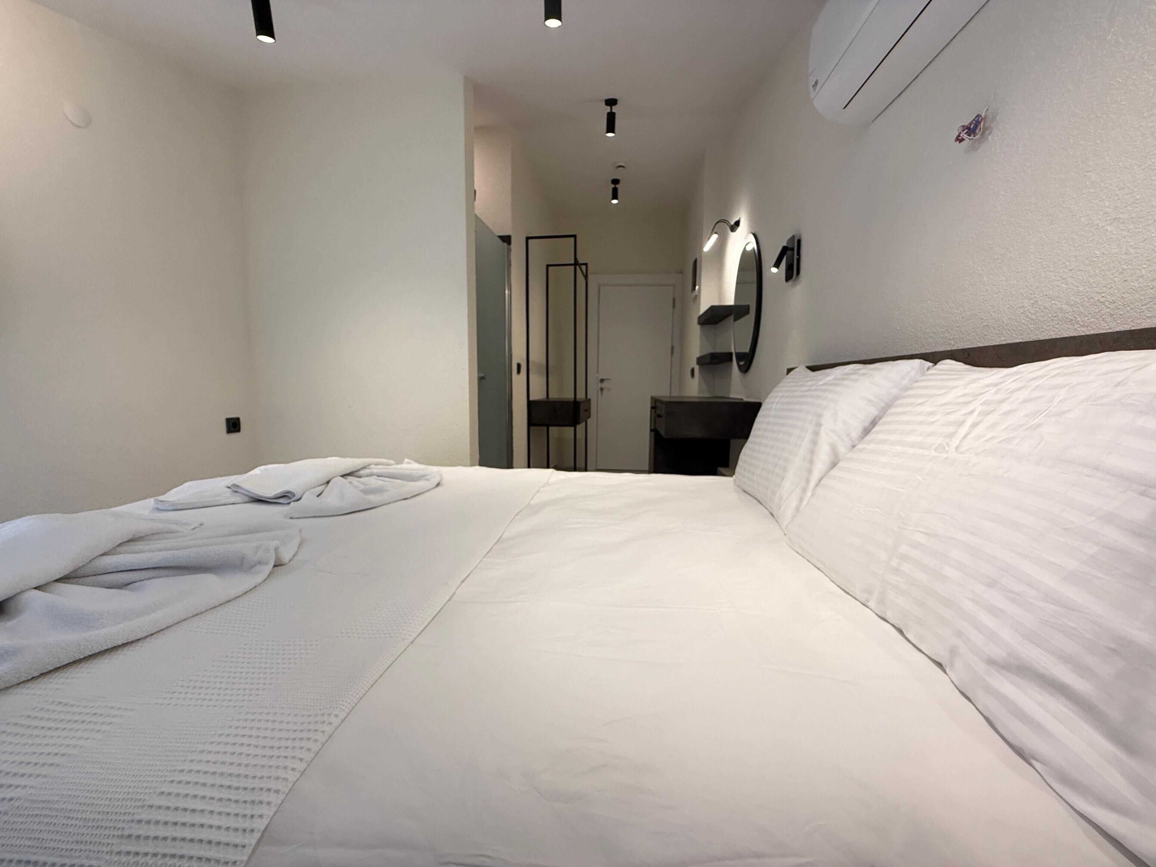 A hotel room with a neatly made bed, a bedside table, a mirror, and a wall-mounted air conditioning unit.