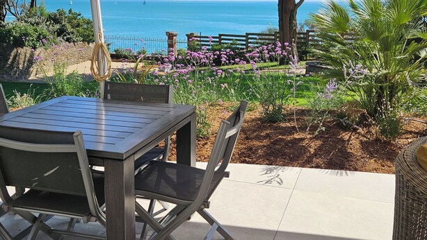 Eine Terrassensitzgelegenheit im Freien mit Tisch und Stühlen, Blick auf das Meer und ein Garten mit violetten Blumen.