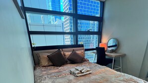 Een slaapkamer met een groot bed, een bureau met lamp en uitzicht op gebouwen door grote ramen.