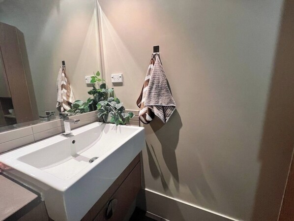 Un baño con un lavamanos blanco, un espejo, un perchero con dos toallas y una planta en maceta.