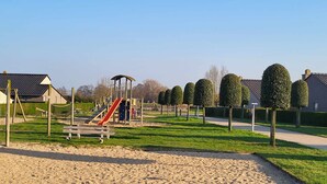 Ein Spielplatz mit Rutsche und Klettergerüsten, umgeben von ordentlich geschnittenen Hecken und blauem Himmel.
