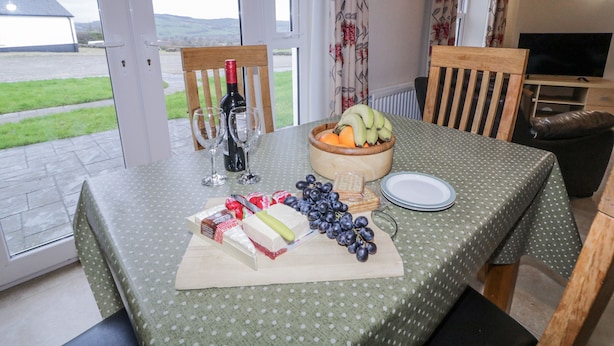 A dining table set with a bottle of wine, glasses, a bowl of fruit, cheese, and grapes. - Foyle Cottage (Strabane)