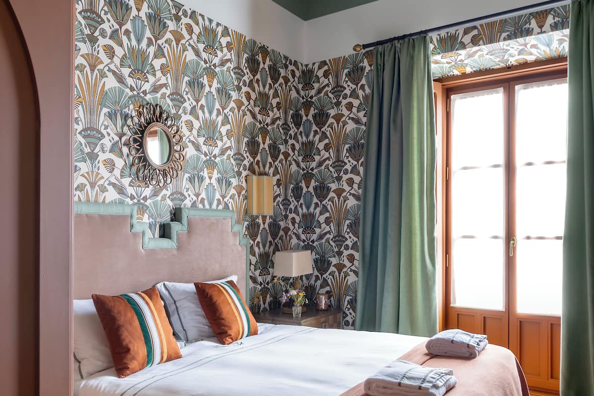 Ein Schlafzimmer mit Bett, Holzboden, gemustertem Wandpapier, einem Spiegel und einem Fenster mit Vorhängen.