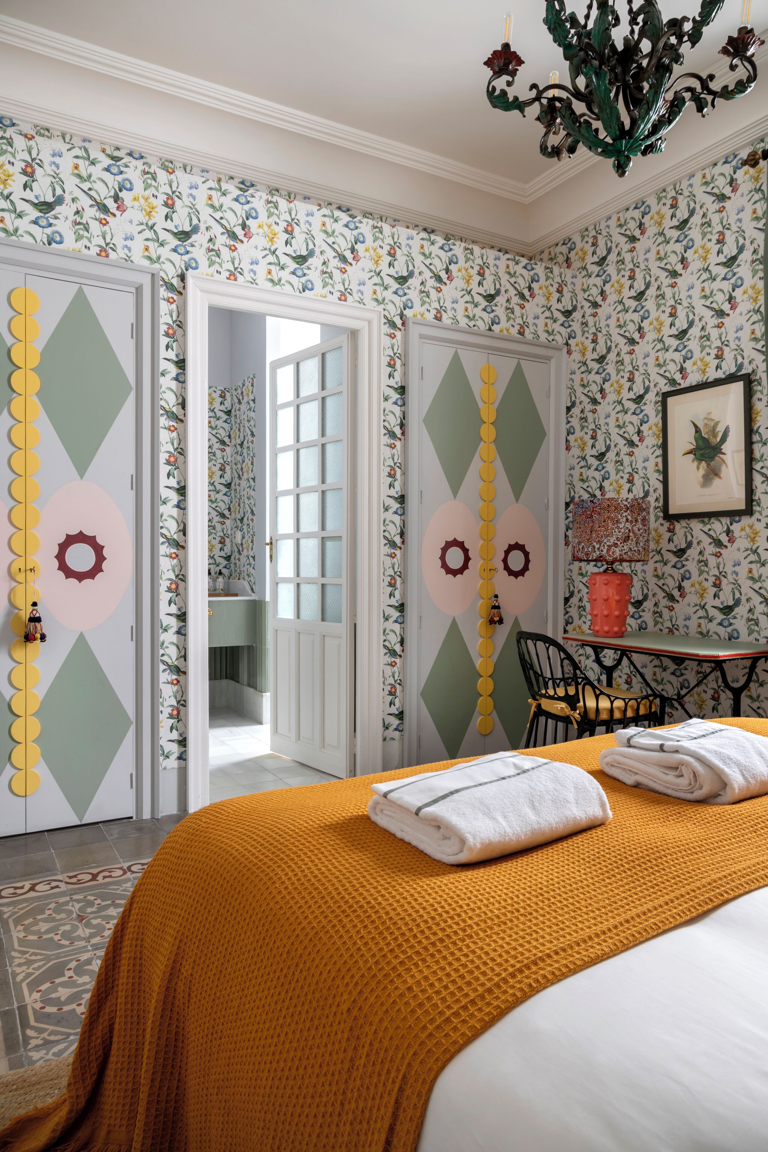 A room with a bed, a chandelier, a door with a geometric pattern, and a table with a chair.