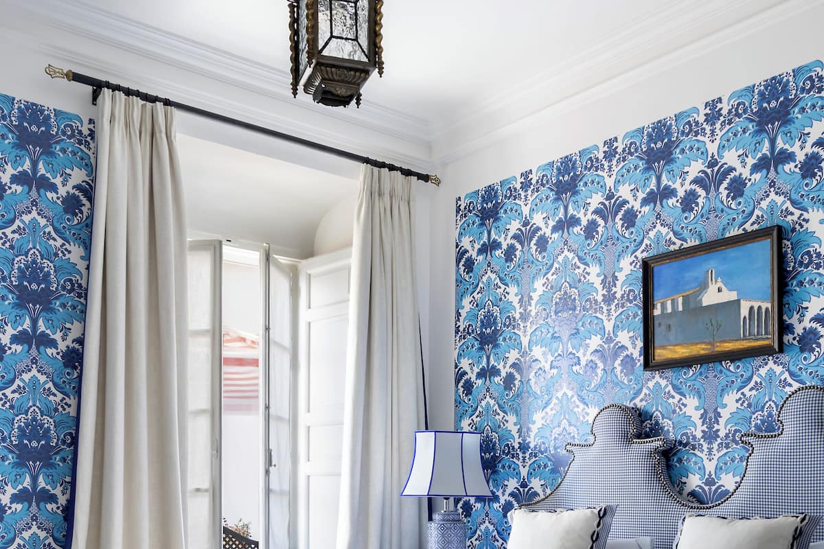 Ein Schlafzimmer mit Bett, Vorhängen, Nachttisch, Lampe und Wand mit blauem Muster.