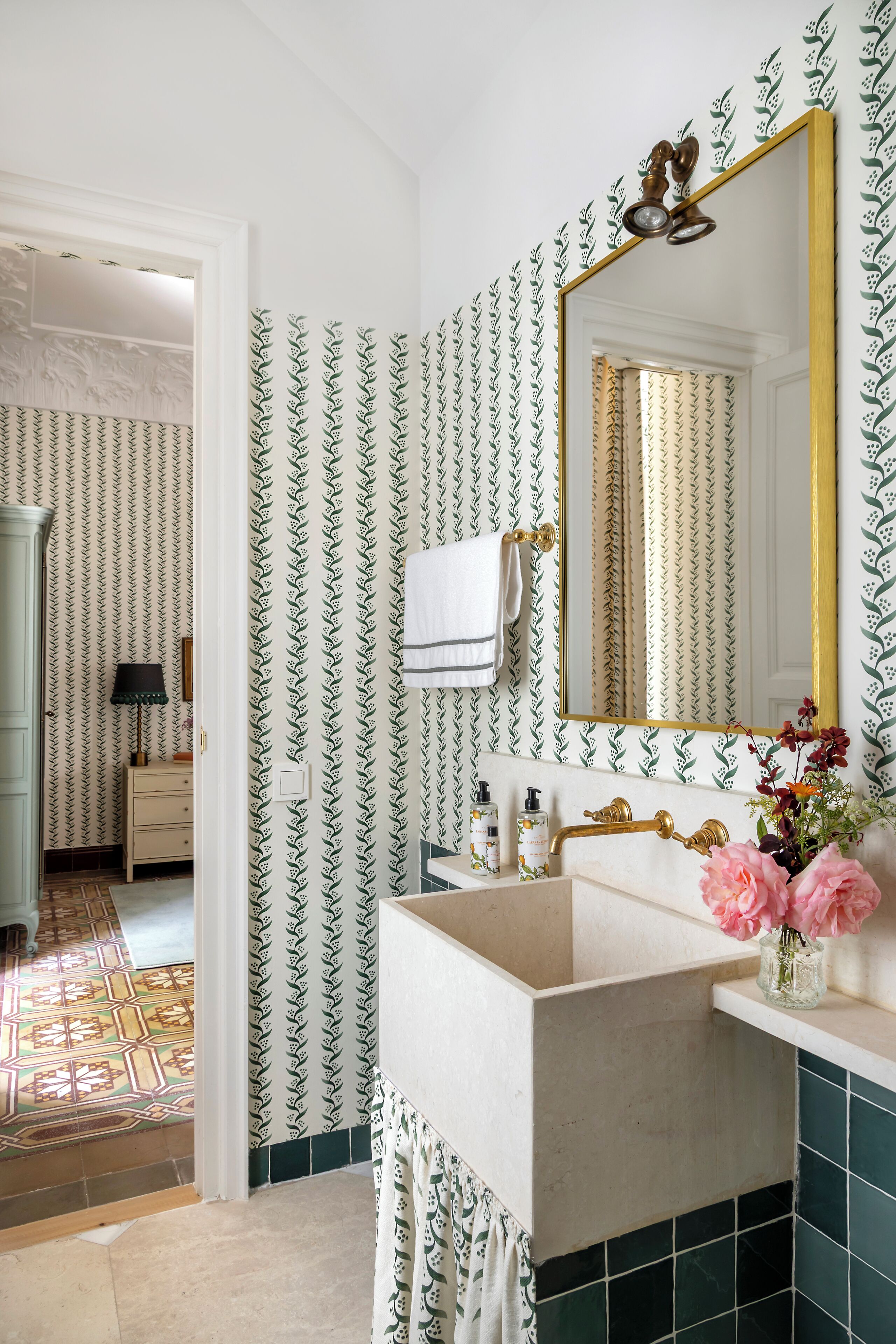 A bathroom with patterned wallpaper, a mirror with a gold frame, a sink with a decorative faucet, and a vase with flowers.