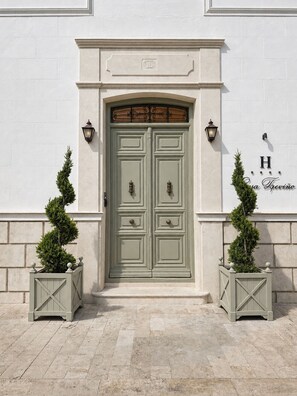 Puertas dobles con un arco decorativo en la parte superior, flanqueadas por dos faroles de pared y dos plantas de hoja perenne en macetas.