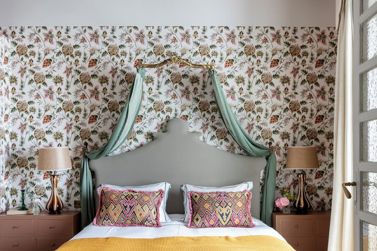 Ein Schlafzimmer mit Baldachinbett, blumigem Tapetenmuster, gefliestem Boden und einem Fenster mit Vorhängen.