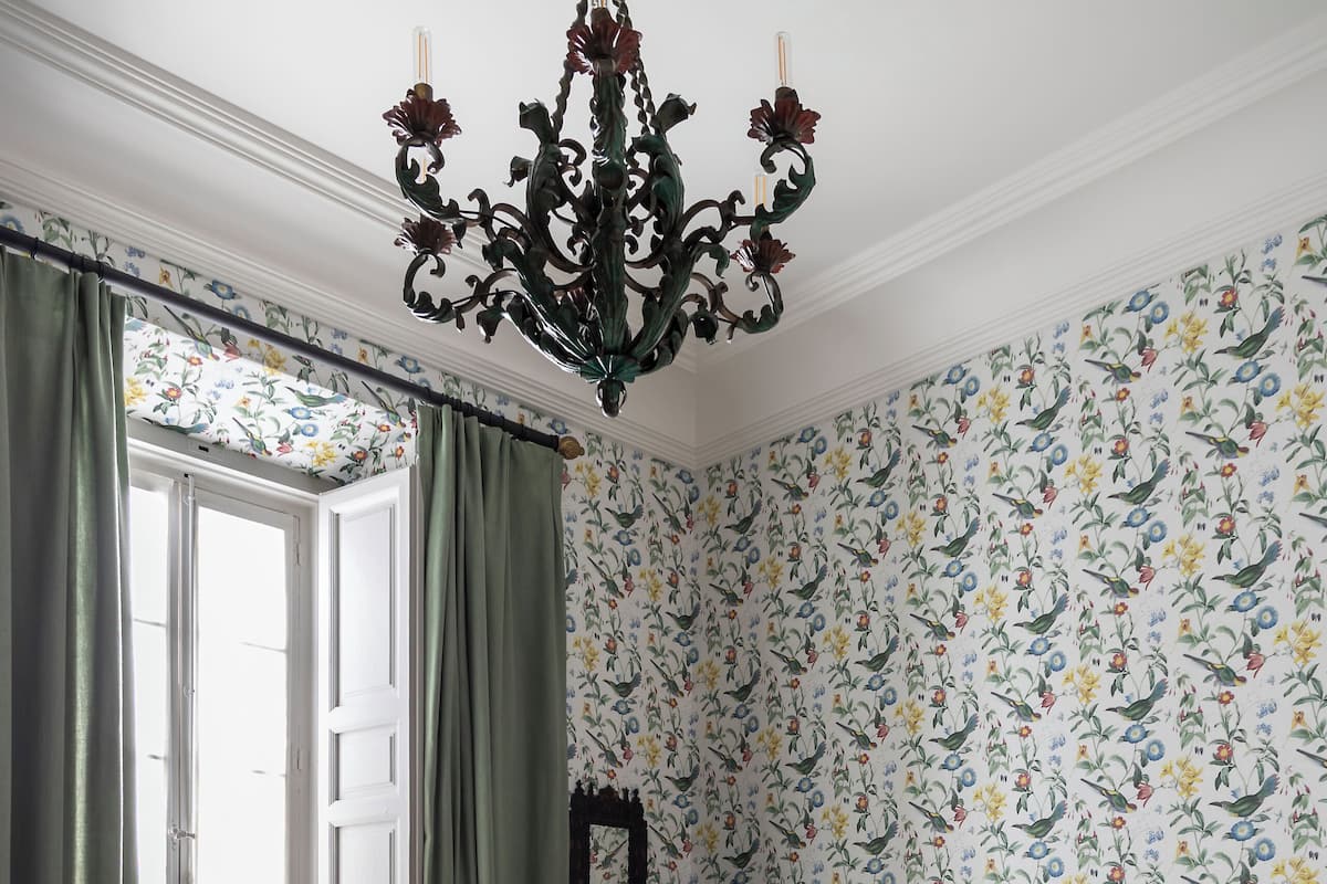 Ein Schlafzimmer mit Bett, Vorhängen, einer Chandelieber, blumigem Wandpapier und einem Fenster.