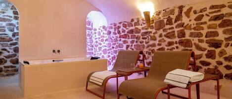 Una habitación con paredes de piedra, una bañera, dos sillones con toallas y una mesa con un vaso.