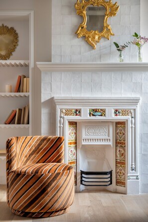 Una habitación con chimenea, un sillón a rayas, una estantería y un espejo decorativo.