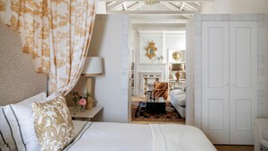 A bedroom with a bed, a nightstand, a lamp, a window with curtains, a wardrobe, and a hallway leading to a bathroom. - Hotel Casa Treviño (Campo de Criptana)