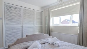 A bedroom with a bed, two rolled towels, a radiator, a window with curtains, and a white ceiling light. - Thorntons House (Northallerton)