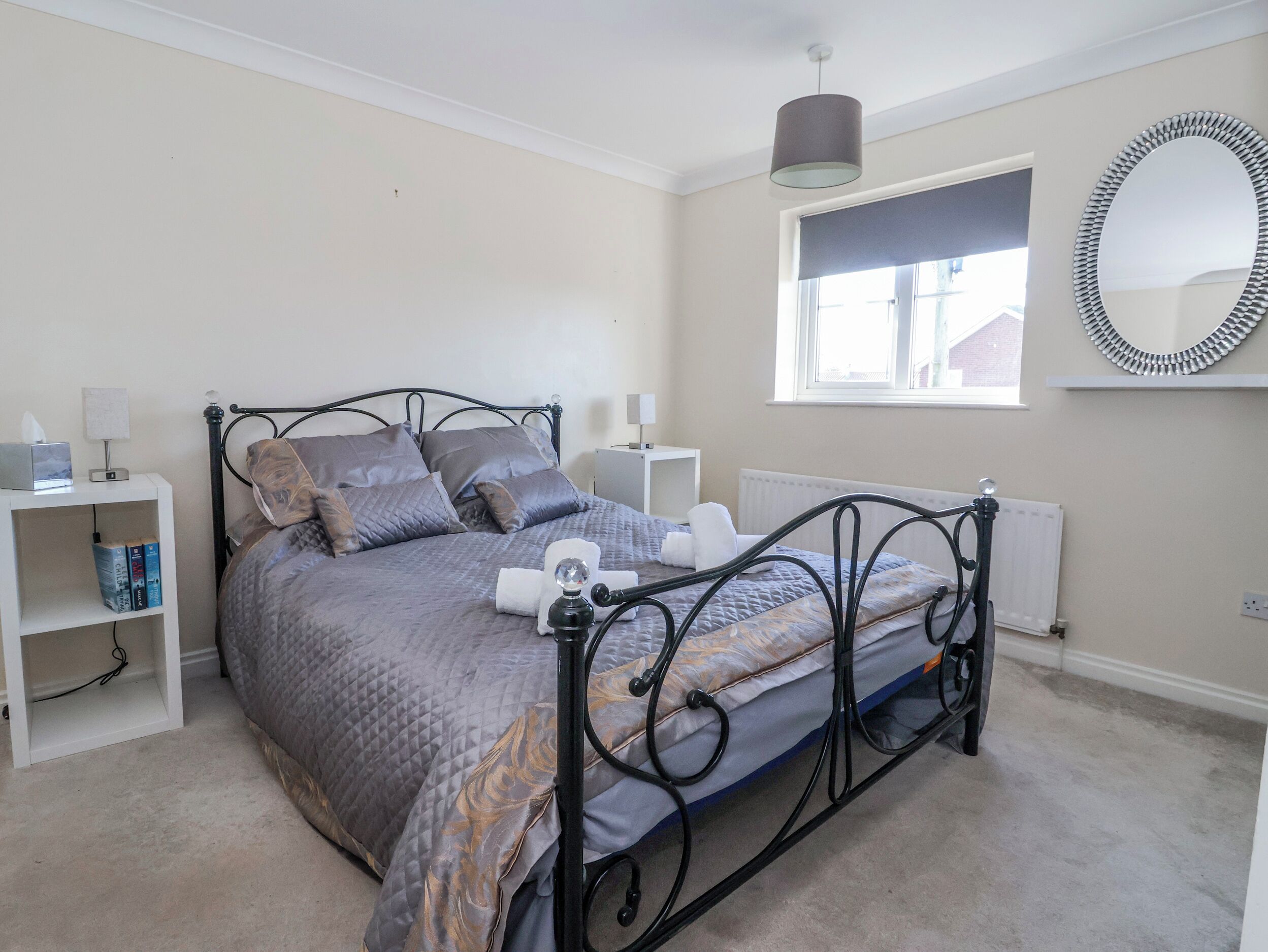 A neatly arranged bedroom with a bed, bedside tables, a lamp, a mirror, and a radiator.