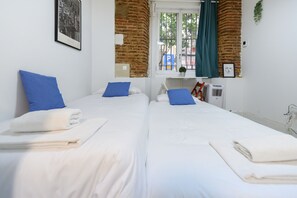 Ein Hotelzimmer mit zwei Betten, weißer Bettwäsche, blauen Kissen, einem Fenster mit Blick und einer Ziegelwand.