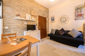 Ein Wohnzimmer mit Holztisch, schwarzem Sofa und Fernseher auf einem weißen Schrank. Eine Tür mit einem 'HOME'-Schild darüber und ein gerahmtes Bild an der Wand.