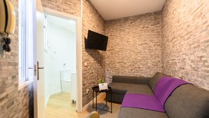 A small, modern room with a brick-patterned wall, a gray sofa with a purple throw, a mounted TV, and a small table with a sign. - M Jpa16bi Acogedor Piso 1 Dormitorio en Tetu n (Madrid)