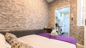 A bedroom with a brick wall, a bed with a purple bedspread, a small table with towels, and a view into the bathroom. - M Jpa16bi Acogedor Piso 1 Dormitorio en Tetu n (Madrid)