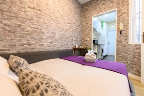 A bedroom with a brick wall, a bed with a purple bedspread, a small table with towels, and a view into the bathroom. - M Jpa16bi Acogedor Piso 1 Dormitorio en Tetu n (Madrid)