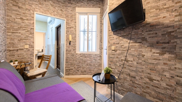A room with a brick wall, a flat-screen TV mounted on the wall, a wooden chair, a small table with a plant, and a purple blanket on the bed. - M Jpa16bi Acogedor Piso 1 Dormitorio en Tetu n (Madrid)