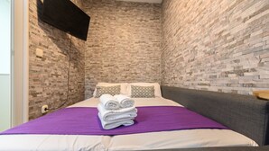A small, modern room with a brick-patterned wall, a bed with a purple blanket, and a flat-screen TV mounted on the wall. - M Jpa16bi Acogedor Piso 1 Dormitorio en Tetu n (Madrid)