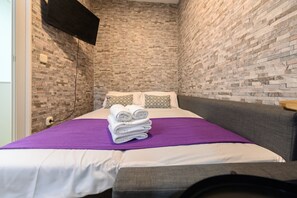 A small, modern room with a brick-patterned wall, a bed with a purple blanket, and a flat-screen TV mounted on the wall. - M Jpa16bi Acogedor Piso 1 Dormitorio en Tetu n (Madrid)