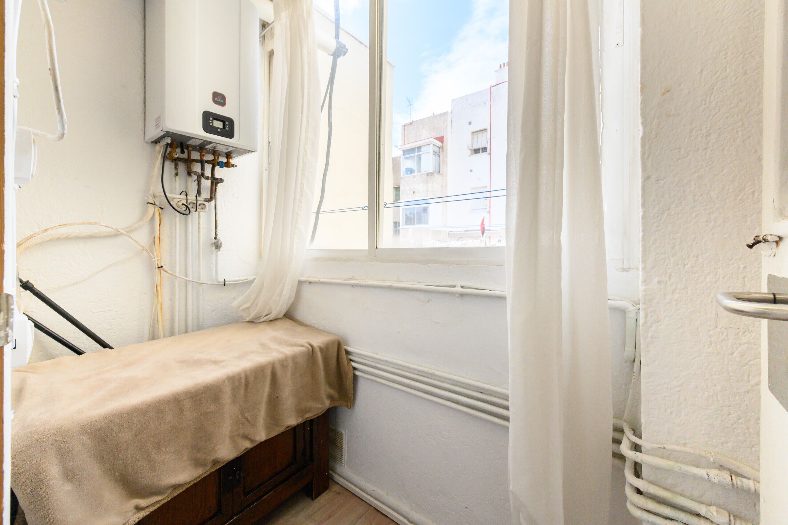 A small, narrow room with a window, a radiator, and a bed with a beige cover.