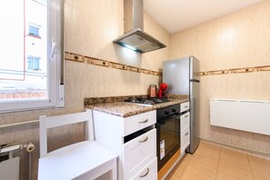 A kitchen with a gas stove, oven, and refrigerator. There is a window with blinds, a chair, and a white cabinet. - M Por183d Amplio Piso 2 Dormitorios en La Latina (Madrid)