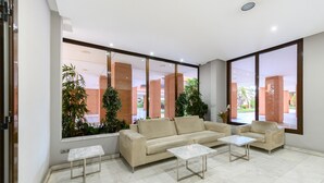 Een ruime lobby met een beige bank, twee witte bijzettafels en grote ramen met uitzicht op een bakstenen gebouw en groen buiten.