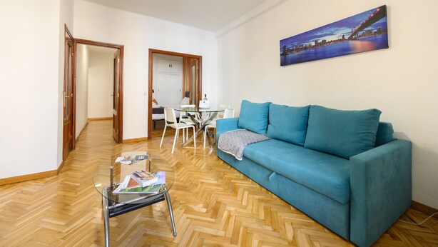 A living room with a blue sofa, a glass coffee table, a dining table with chairs, and a painting on the wall. - M May613 Piso de 1 Dormitorio en Pleno Centro de (Madrid)