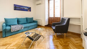 A living room with a blue sofa, a black armchair, a glass coffee table, and a wooden herringbone floor. - M May613 Piso de 1 Dormitorio en Pleno Centro de (Madrid)