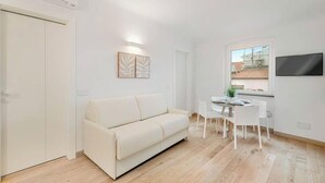 Una sala de estar moderna con un sofá blanco, una mesa de comedor con sillas, un televisor de pantalla plana montado en la pared y un ventanal con vistas al exterior.