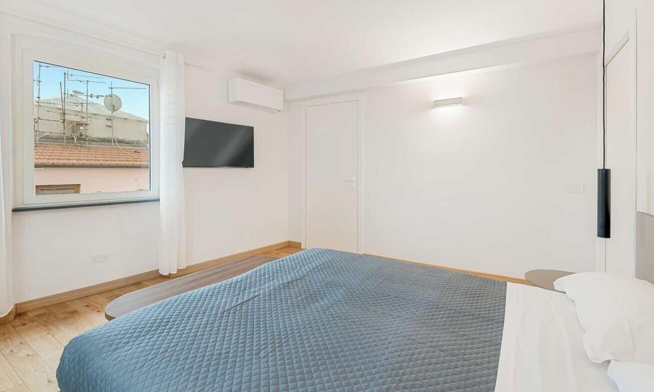 Ein modernes Schlafzimmer mit einem Bett, einem an der Wand befestigten Fernseher und einem Fenster mit Blick auf Gebäude.