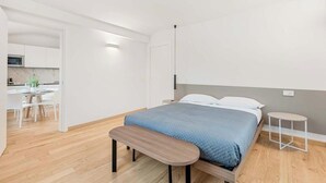Un dormitorio moderno con cama, banqueta, mesita de noche y vista a una zona de cocina.