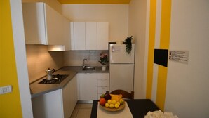 Cozinha compacta com armários brancos, pia, fogão e geladeira. Há uma mesa pequena com uma fruteira e um vaso de planta.
