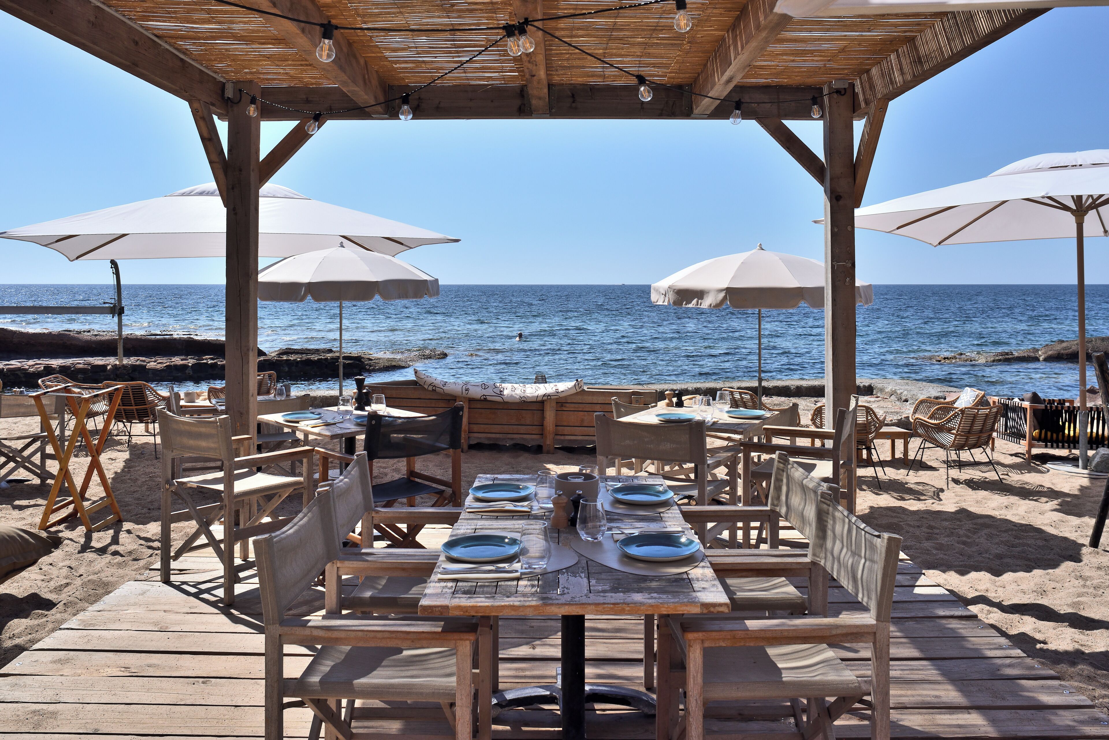 A beachside restaurant with wooden tables and chairs, umbrellas, and a view of the sea.