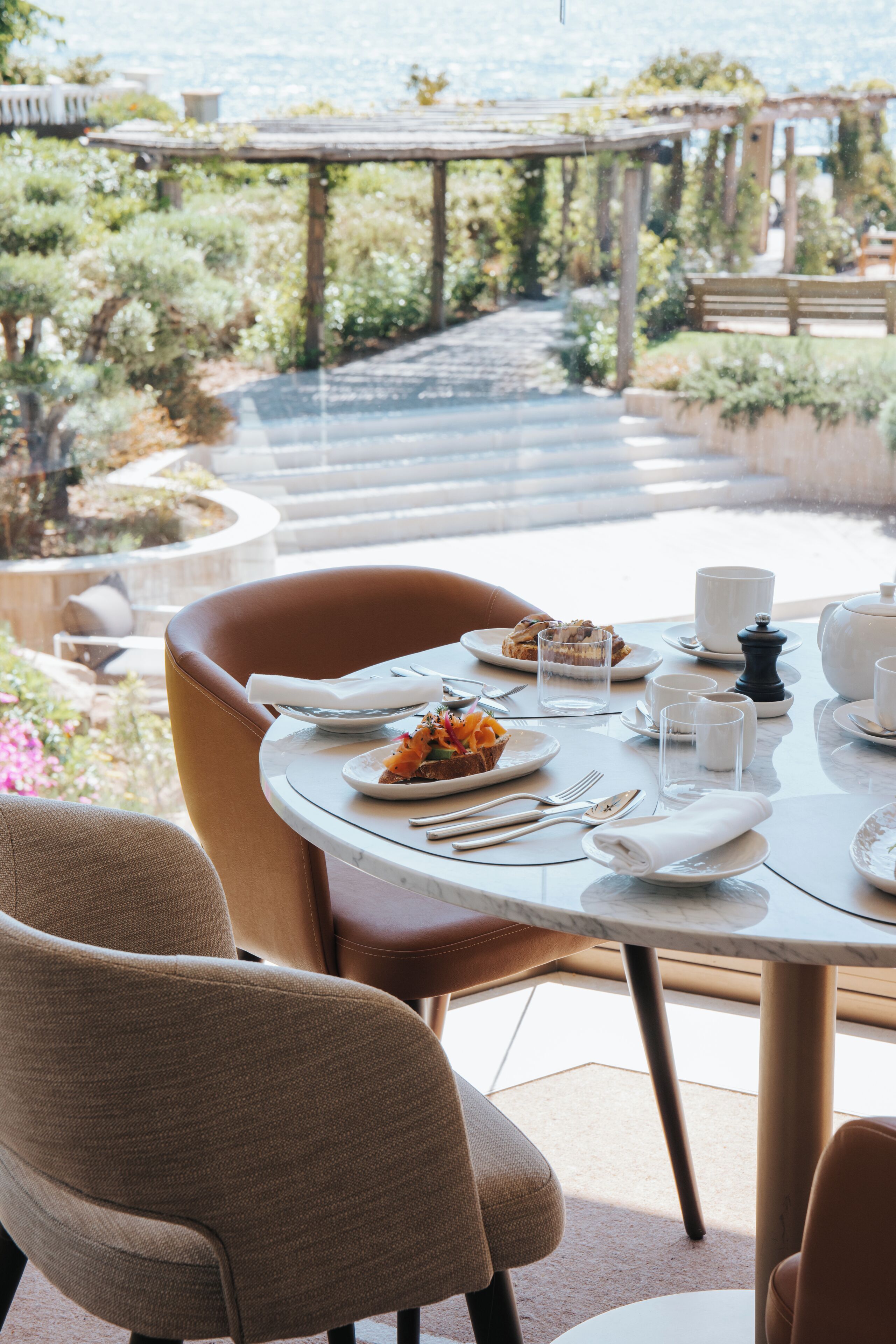 A dining table set with plates, glasses, and cutlery, overlooking a garden with a wooden pergola.