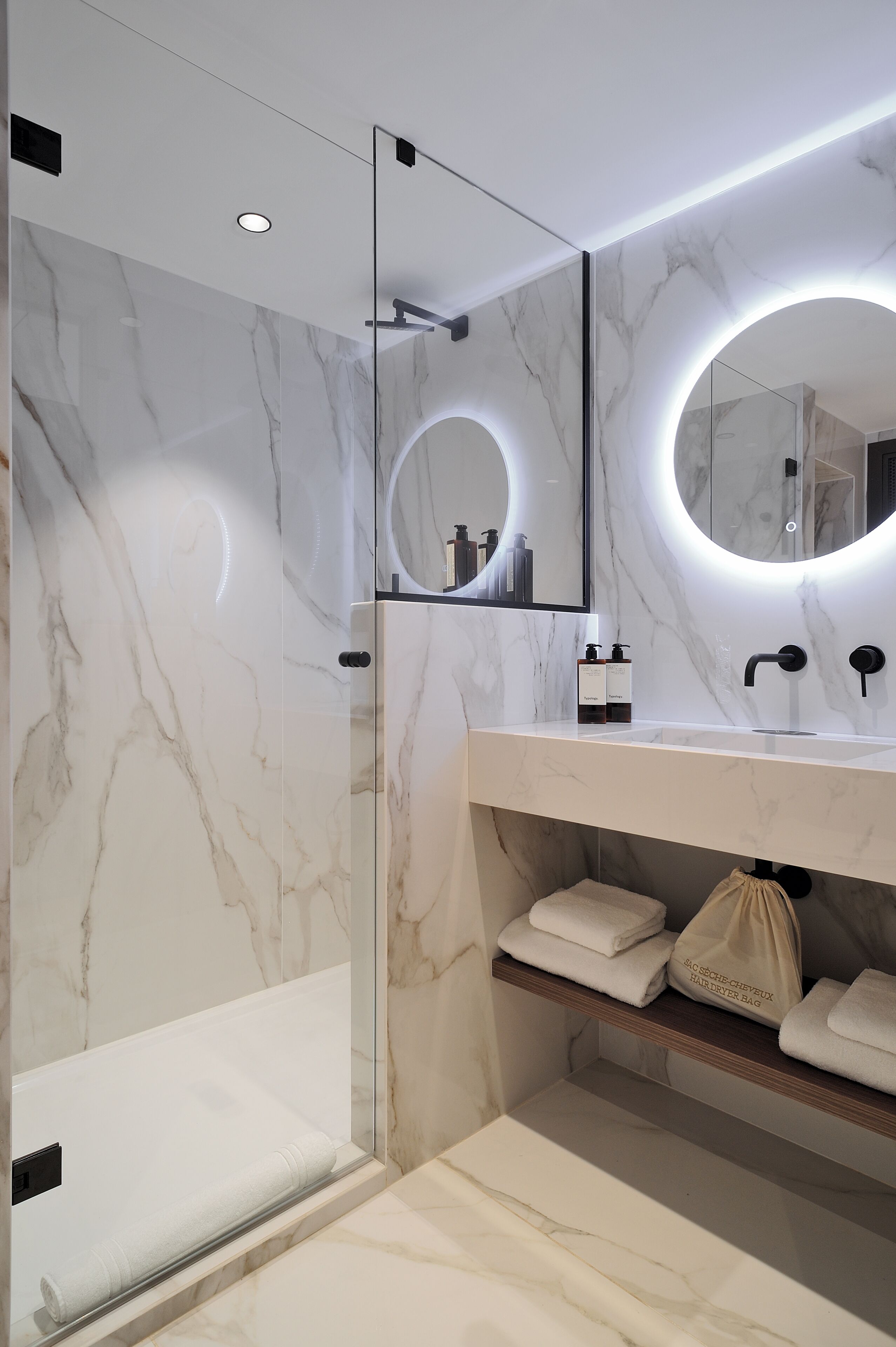 Un baño moderno con ducha de vidrio, tocador de mármol y espejo con luz circular.