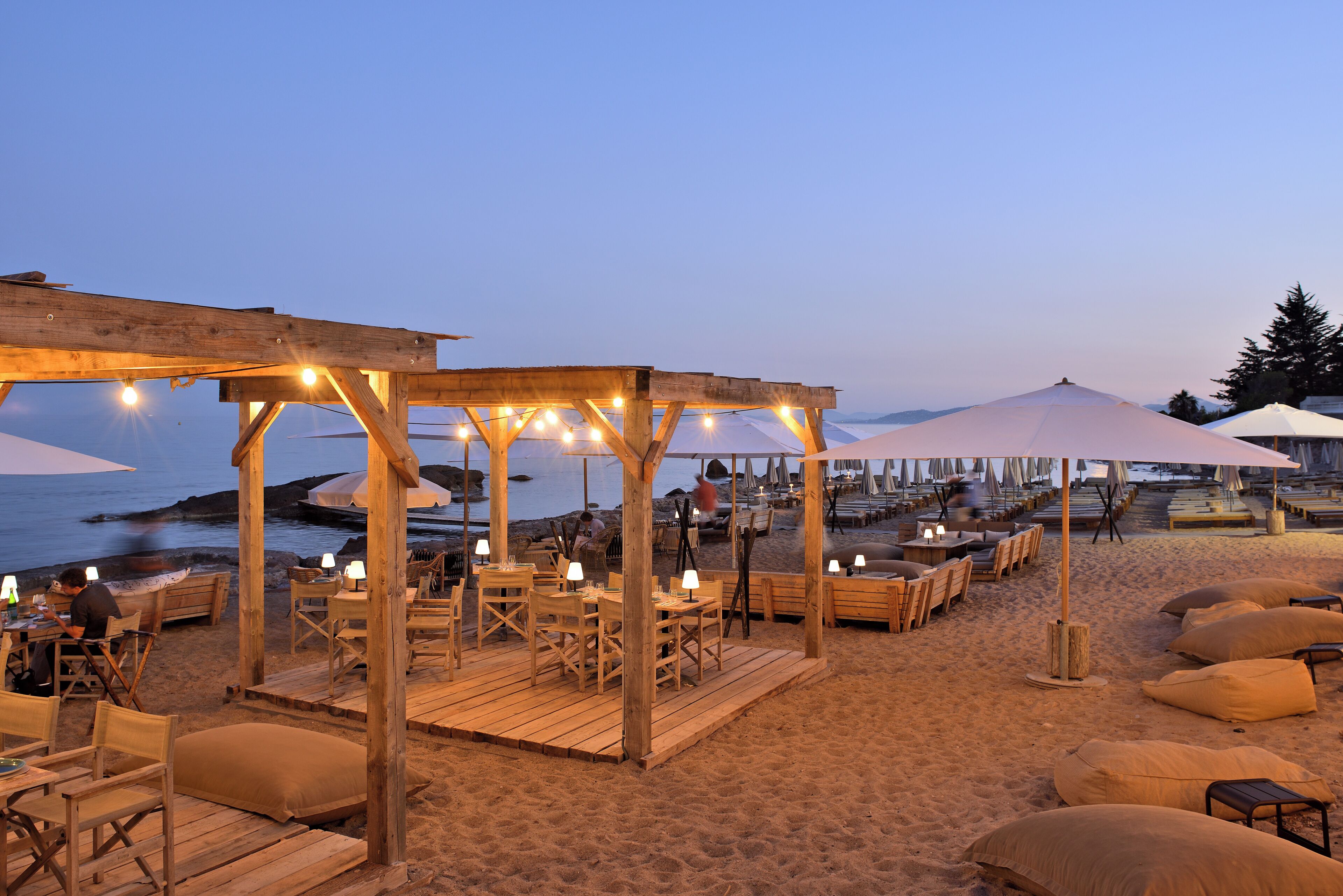 A beachside restaurant with outdoor seating, tables, and umbrellas.