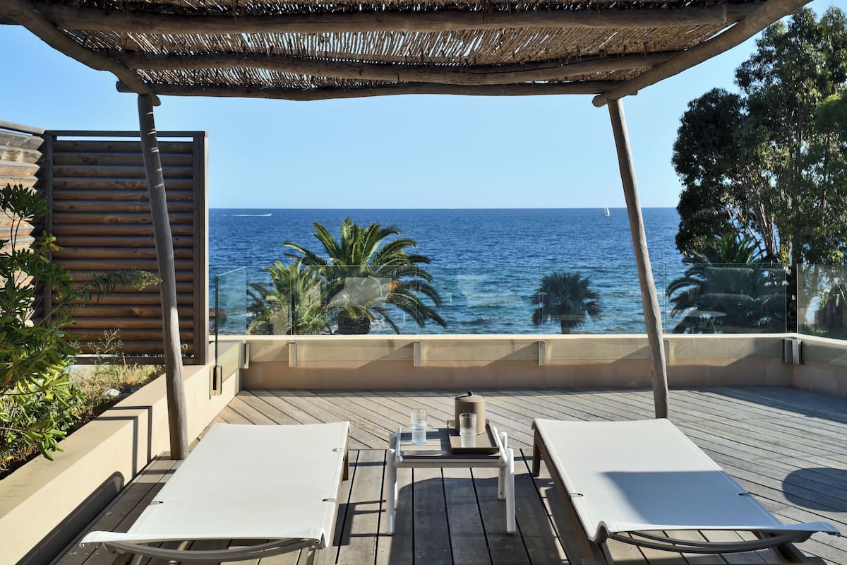 Eine Holzterrasse mit zwei weißen Liegestühlen und einem Tisch, mit Blick auf das Meer und freier Sicht auf das offene Meer und ein Segelboot.