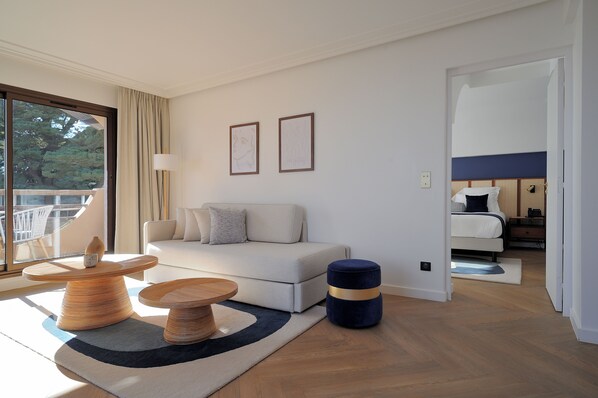 A modern hotel room with a sofa, two framed artworks, a round wooden table, a blue ottoman, and a view of the outdoors. - La Réserve Esterel (Saint-Raphaël)