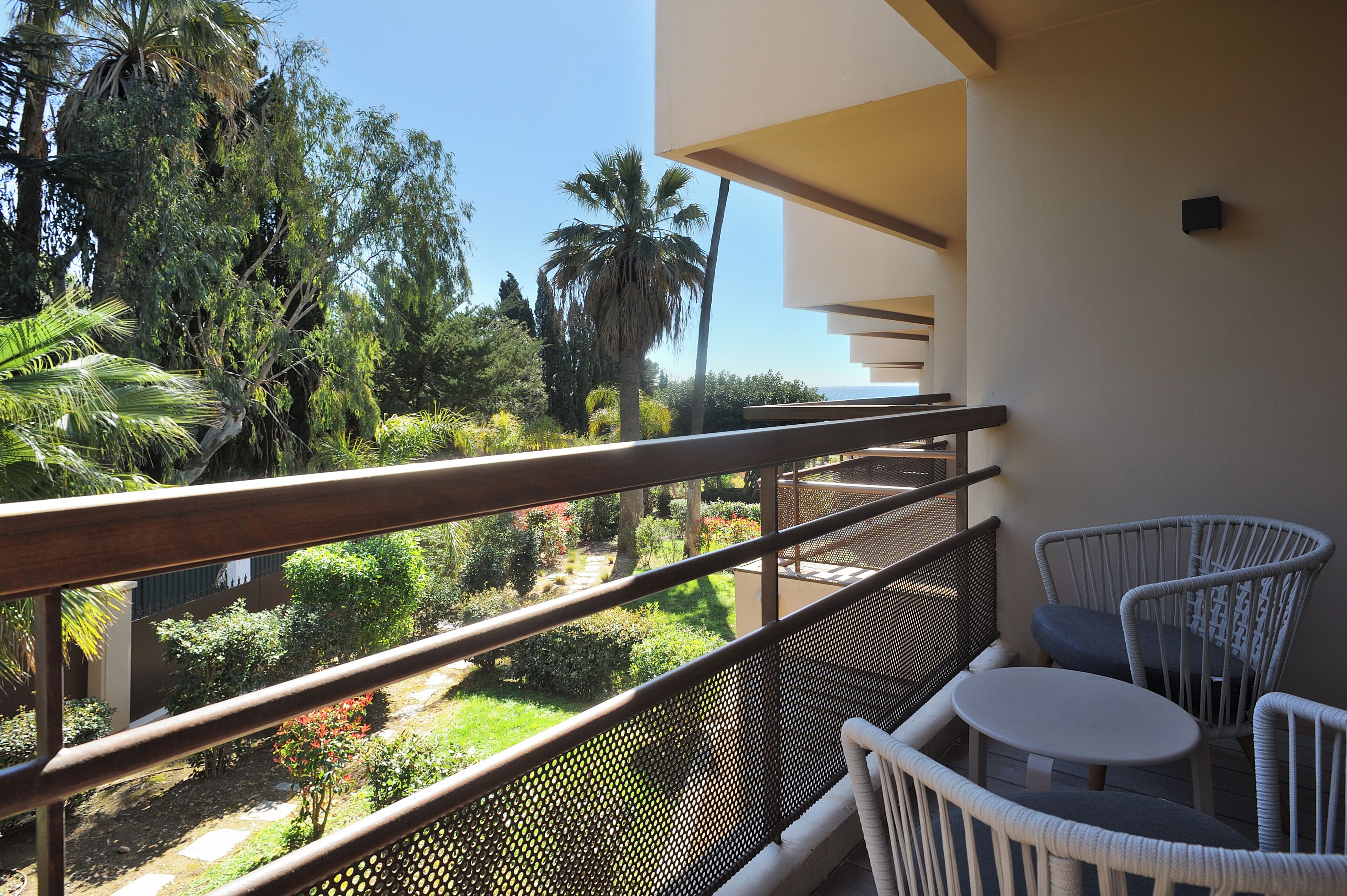 A balcony with a white table and chairs, overlooking a garden with various plants and trees.