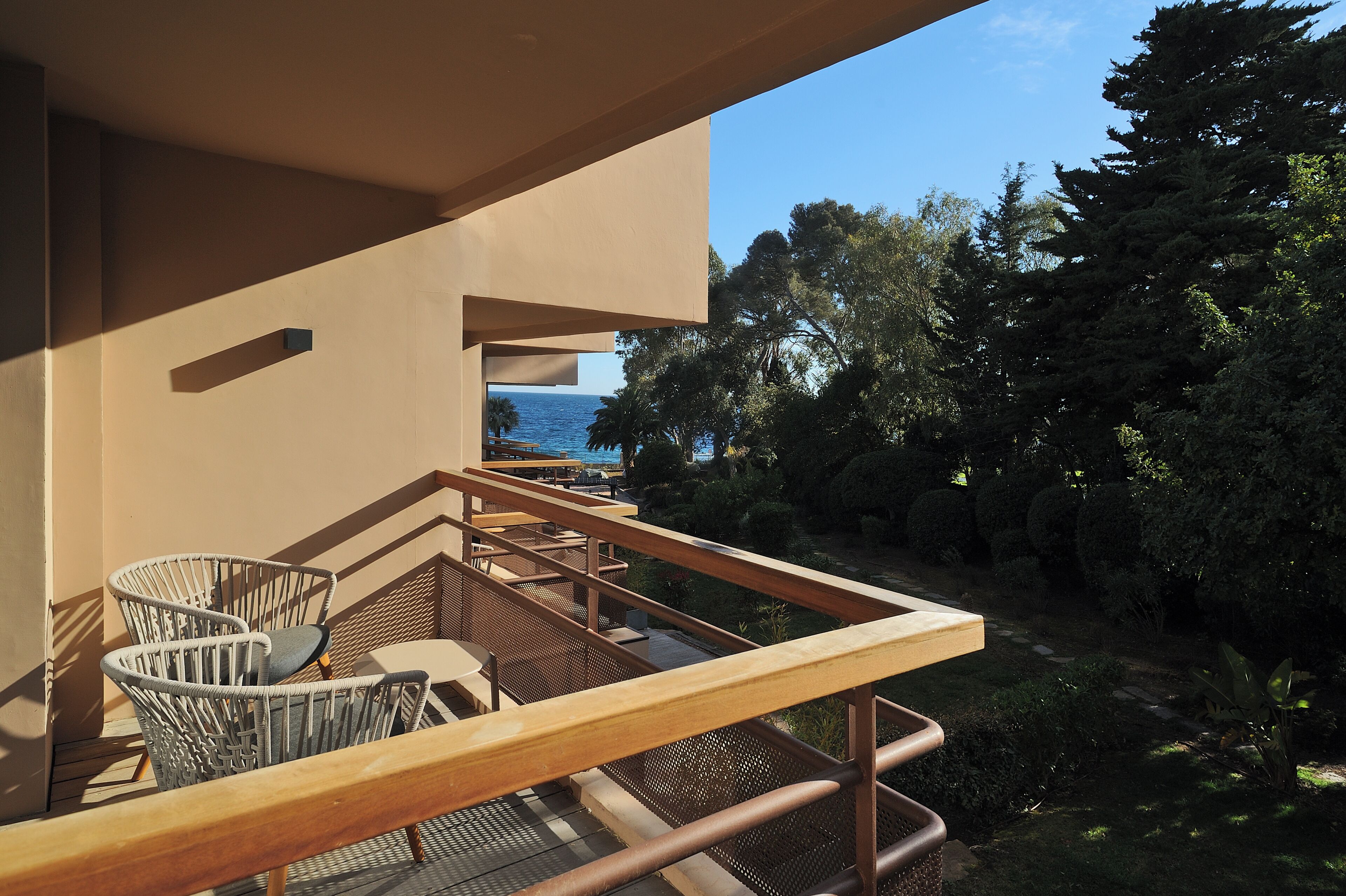 Un balcón con sillas de mimbre y una mesa, con vista a un jardín y una masa de agua.