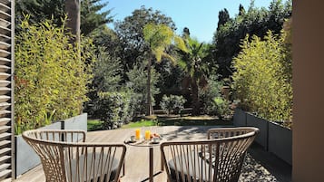 Un patio in legno con sedie in vimini, un tavolo imbandito per la colazione e vista su un giardino con diversi alberi e una palma.