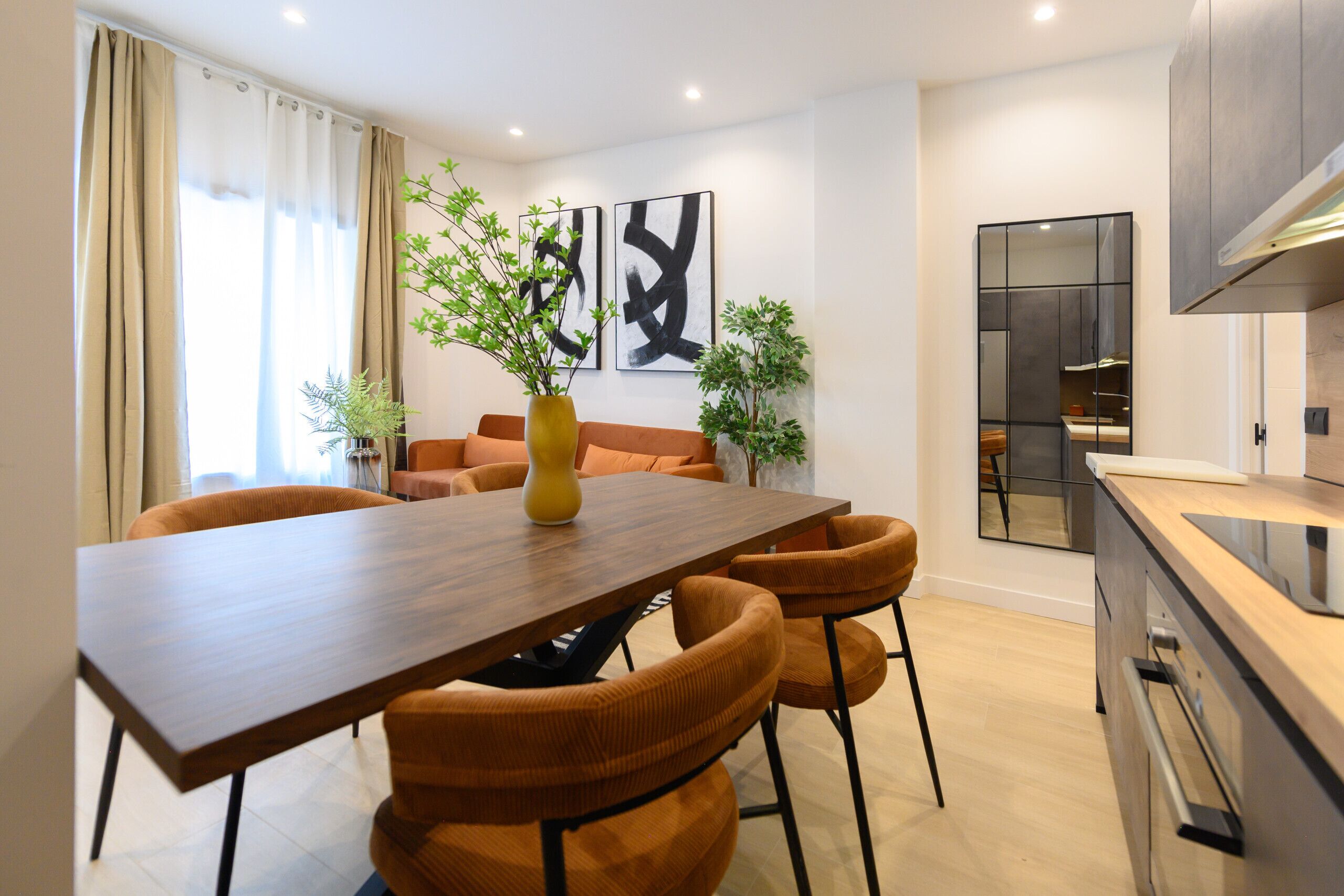 A modern kitchen with a wooden table and chairs, a built-in oven, and a large window with curtains.