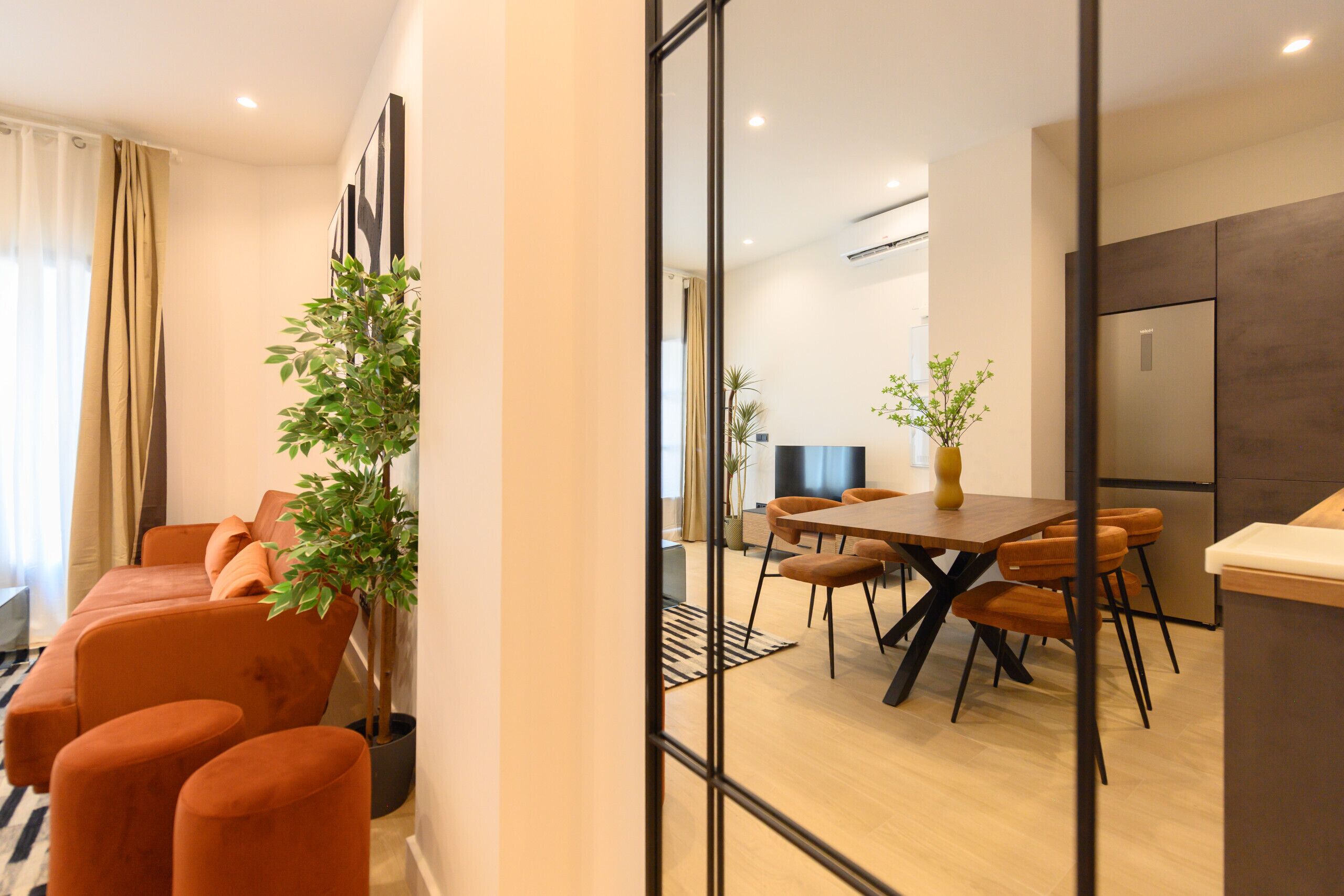 A modern living room with orange sofas, a dining area with a wooden table and chairs, and a kitchen with a refrigerator.