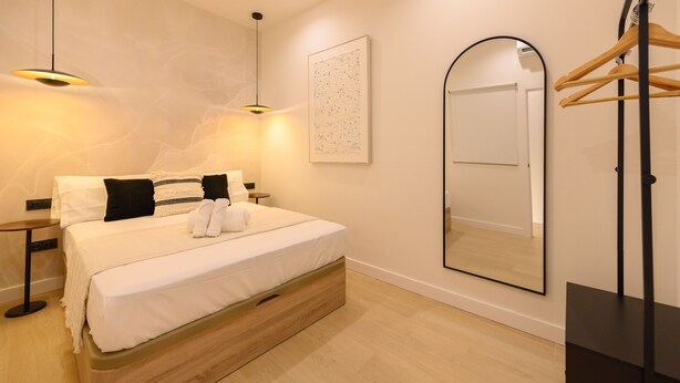 A neatly made bed with white linens and black pillows, a bedside table with a lamp, a large mirror, and a wooden hanger rack. - M Agu230 Tu Hogar en la Ciudad Piso de 3 Habita (Madrid)