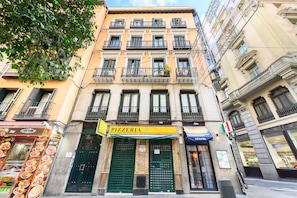 Un immeuble dont le rez-de-chaussée abrite une pizzeria, reconnaissable à son auvent vert et à une enseigne portant le mot « PIZZERIA ».